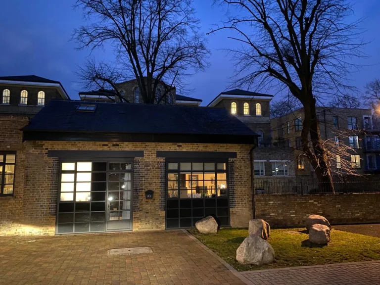 Twilight view of a modern brick building with large windows and extensions, flanked by trees and decorative boulders, against a backdrop of traditional structures. Loft Conversions, House Extensions, Roofing, Bespoke Loft Conversions, House Renovations